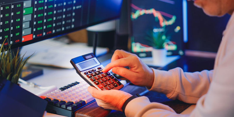 A person working through figures on a calculator in front of screens of financial data