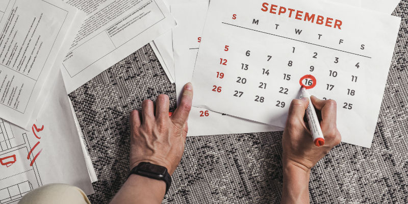 A hand circling a date on a paper calendar laid over stacks of corporate paperwork